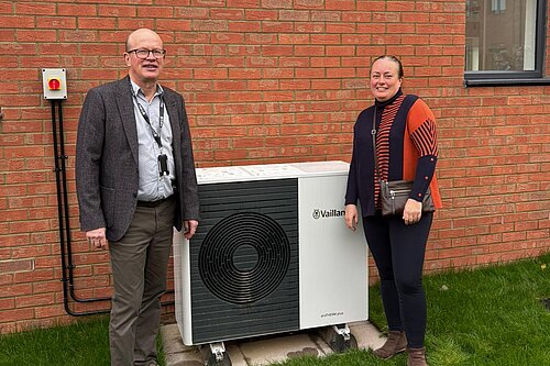 Cllr Paul de Kort and Cllr Jacqui Taylor are standing on either side of the air source heat pumps at the new properties. They are both smiling and looking at the camera, and are stood in front of the brick wall of one of the new properties. 